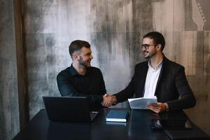 dos empresarios en un oficina sonriente a el cámara mientras trabajando juntos detrás un ordenador portátil computadora. foto