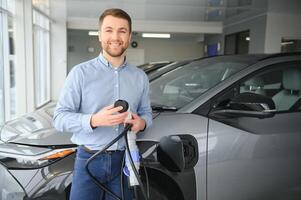 Handsome business man holding charging cable for electric car. Caucasian male stands near electric auto in dealership photo