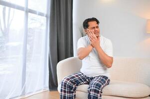 Dental problems. Young indian man touching cheek, closing eyes with expression of terrible suffer from painful toothache, sensitive teeth, cavities. Hindu guy at home apartment living room on sofa photo