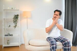 Dental problems. Young indian man touching cheek, closing eyes with expression of terrible suffer from painful toothache, sensitive teeth, cavities. Hindu guy at home apartment living room on sofa photo