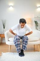 Unhappy indian or arabian man, sits on comfortable sofa in cozy living room, holds his hands on his stomach, grimaces from pain in his stomach, suffers from poisoning, spasm, stomach problems. photo