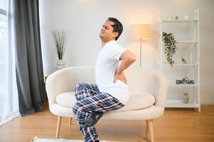 A sick man on Christmas has severe back pain, massages muscle his hand sitting on sofa in living room photo