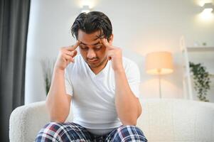 retrato de joven indio chico sufrimiento desde migraña a hogar, oriental hombre sensación indispuesto, conmovedor su templos con cerrado ojos, sufrimiento agudo dolor de cabeza mientras sentado en sofá foto