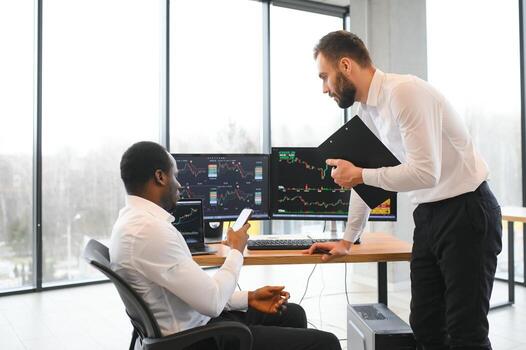 Two diverse colleagues traders talking to each other, sitting in the office in front of multiple computer screens. Stock trading, people, business concept photo
