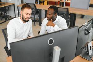 Two diverse colleagues traders talking to each other, sitting in the office in front of multiple computer screens. Stock trading, people, business concept photo