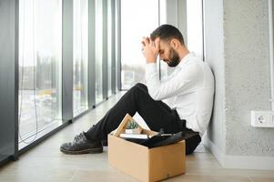 joven hermoso empresario en ligero moderno oficina con caja de cartón caja. último día a trabajar. trastornado oficina trabajador es despedido foto