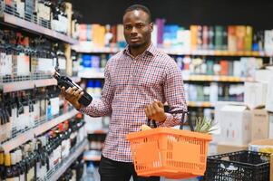 African American man holding bottle of wine and looking at it while standing in a wine store photo