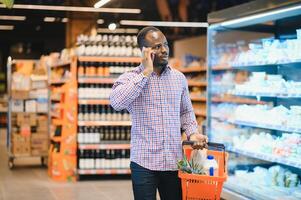 africano americano hombre hablando en Teléfono móvil chateando mientras compras comestibles en supermercado foto