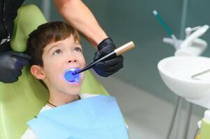 un pequeño chico teniendo su diente hecho en el clínica - poniendo el fotopolímero lámpara con azul ligero en el boca foto
