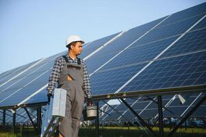 Portrait of Young indian man technician wearing white hard hat standing near solar panels against blue sky. Industrial worker solar system installation photo
