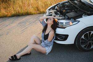 Beautiful young girl sitting at a broken car on the road desperate to get help photo