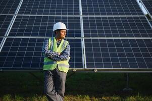 retrato de joven indio hombre técnico vistiendo blanco difícil sombrero en pie cerca solar paneles en contra azul cielo. industrial trabajador solar sistema instalación foto