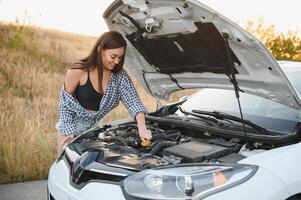 Beautiful slim girl in shirt and shorts looks in open car hood on a road photo