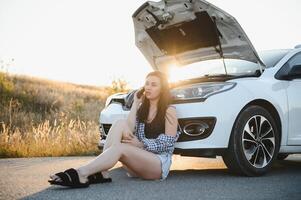 Beautiful young girl sitting at a broken car on the road desperate to get help photo