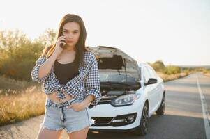 el joven niña rompió el coche y ella abrió el capucha y intentó a reparar el coche en el la carretera. un mujer llamadas en un teléfono inteligente con un coche evacuación Servicio foto