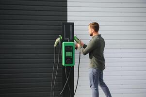 un hombre soportes cerca un cargando estación y cargos su eléctrico coche foto