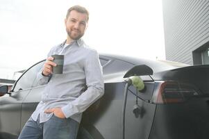 handsome bearded man in casual wear, standing at the charging station and charger for an electric car. Eco electric car concept photo