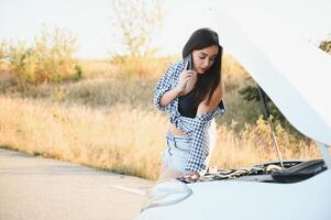 atractivo Delgado joven niña en verano pantalones cortos y camisa refacción un roto coche. un hermosa mujer soportes cerca elevado coche capucha. foto