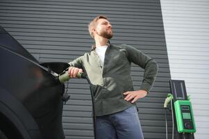 handsome bearded man in casual wear, standing at the charging station and charger for an electric car. Eco electric car concept photo