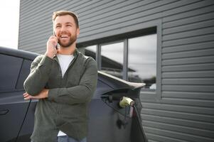 Happy man using smart phone and charging car at vehicle charging station photo