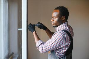 africano americano obrero en mono instalando o ajustando el plastico ventanas en el vivo habitación a hogar foto