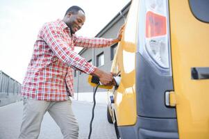 African american Man stands next to electric delivery vans at electric vehicle charging station photo