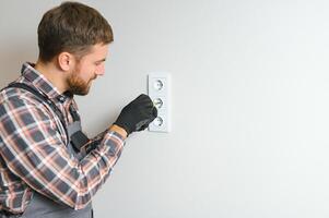 confidente profesional electricista en uniforme utilizando destornillador mientras reemplazando un enchufe en Departamento después renovación trabajar. foto