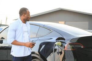 elegante africano hombre con café taza en mano inserta enchufe dentro el eléctrico coche cargando enchufe foto