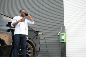 Stylish african man with coffe cup in hand inserts plug into the electric car charging socket photo