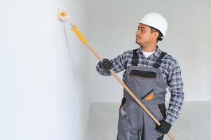 un indio Departamento reparar trabajador pinturas un blanco pared con un rodillo. foto