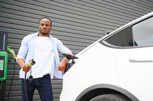 African man standing near electric car with charging cable in plug. Eco friendly vehicle charging on station. LIfestyles concept photo