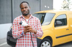 africano americano hombre soportes siguiente a eléctrico entrega furgonetas a eléctrico vehículo cargando estación foto