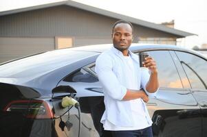 African man standing near electric car with charging cable in plug. Eco friendly vehicle charging on station. LIfestyles concept photo