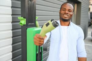 Serious african man holding charge cable in on hand, standing near luxury electric car. photo