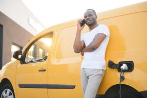 African american Man stands next to electric delivery vans at electric vehicle charging station photo