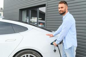 hermosa joven elegante hombre es con eléctrico coche a tiempo de día cargando el vehículo foto