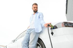 Beautiful young stylish man is with electric car at daytime charging the vehicle photo