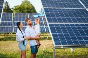 un amplio Disparo de un contento familia en pie juntos y sonriente a cámara con un grande solar panel en antecedentes foto