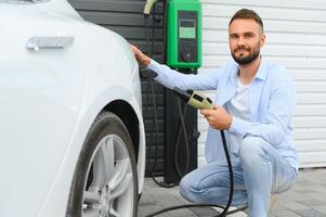 Beautiful young stylish man is with electric car at daytime charging the vehicle photo