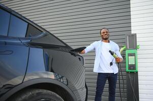 Serious african man holding charge cable in on hand, standing near luxury electric car. photo