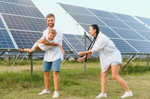 joven familia de Tres es agachado cerca fotovoltaica solar panel, pequeño chico y padres. moderno familia concepto. el concepto de verde energía foto