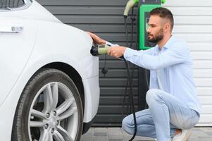 Beautiful young stylish man is with electric car at daytime charging the vehicle photo