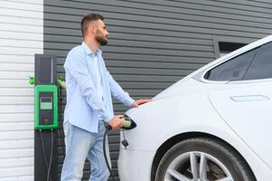 Beautiful young stylish man is with electric car at daytime charging the vehicle photo