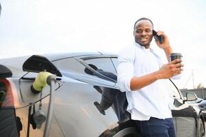 elegante africano hombre con café taza en mano inserta enchufe dentro el eléctrico coche cargando enchufe foto