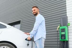 Beautiful young stylish man is with electric car at daytime charging the vehicle photo