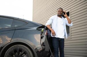 African man standing near electric car with charging cable in plug. Eco friendly vehicle charging on station. LIfestyles concept photo