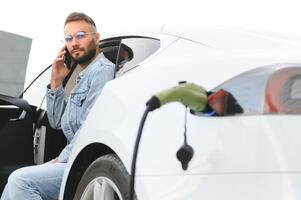 joven elegante hombre es con eléctrico coche a tiempo de día. el concepto de verde energía foto