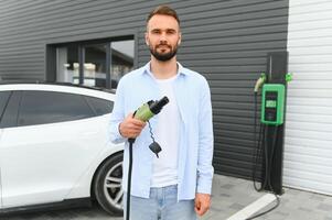 joven hermoso hombre participación cargando cable a eléctrico cargando estación punto en pie cerca su nuevo coche foto