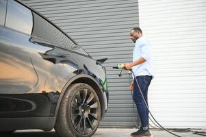 Serious african man holding charge cable in on hand, standing near luxury electric car. photo