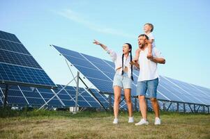 joven familia de Tres es agachado cerca fotovoltaica solar panel, pequeño chico y padres. moderno familia concepto. el concepto de verde energía foto